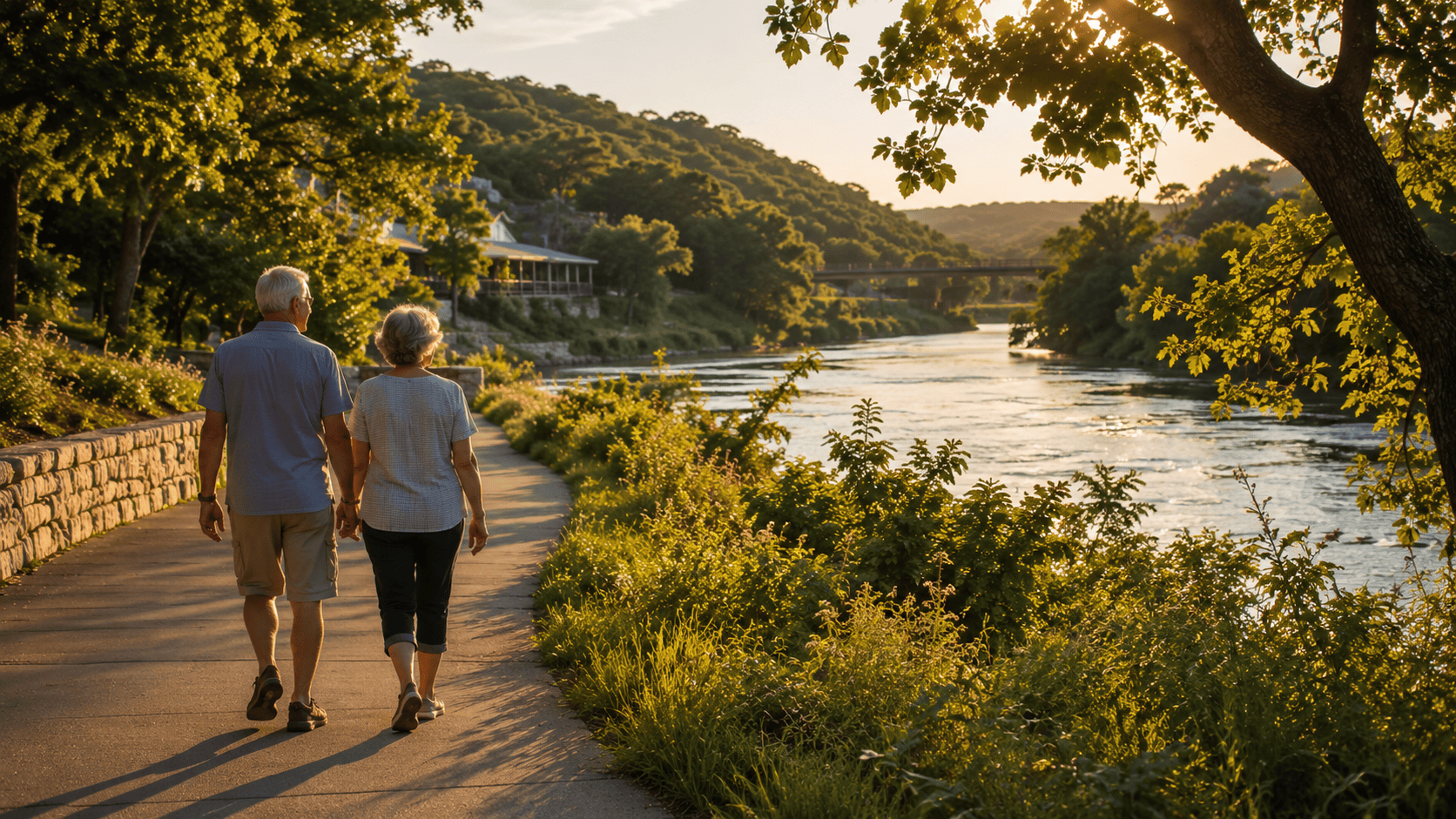 A retired couple walks along the Comal River in New Braunfels, Texas on a sunny morning, with cypress trees lining the clear water and a Hill Country backdrop